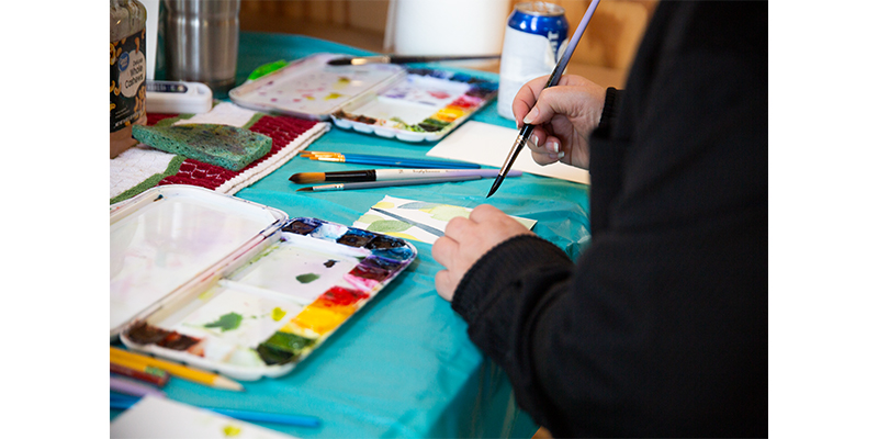 A hand with a paintbrushes painting a small canvas in watercolor