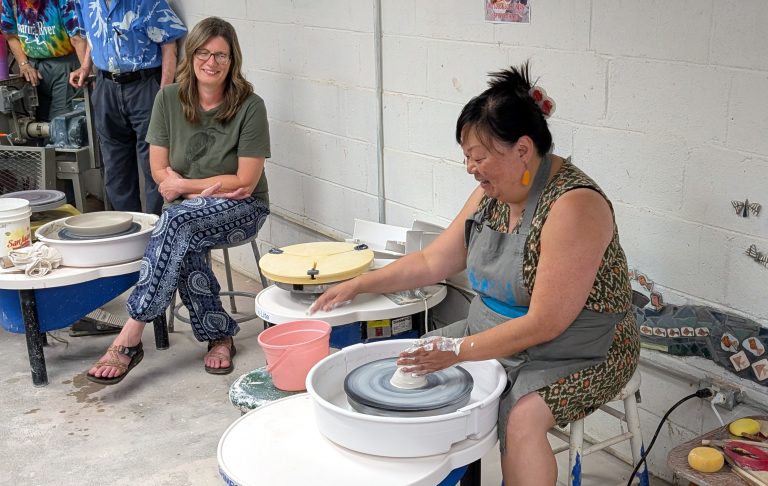 Two women sit in front of ceramics wheels smiling. The woman closest to the camera is actively making pottery on the wheel
