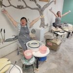 Two women sitting in front of clay throwing wheels with their arms in the air and huge smiles on their face