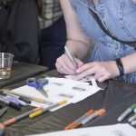 Person drawing designs on paper at a table covered with markers, scissors, and art supplies.
