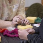 Hands shaping wool with a felting needle over a foam block during a fiber arts activity.