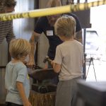 Two children watch as an instructor guides them hammering heated metal on an anvil in a blacksmithing studio.