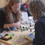 An instructor wearing safety glasses guides a participant in soldering stained glass pieces at a workbench.