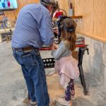 An adult instructor and a small child wearing face shields work together at a wood lathe, shaping wood in a workshop.