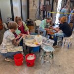 Several people sit at pottery wheels in a studio, shaping clay pieces together with tools and buckets nearby.