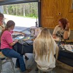 A group of participants sits around a table creating custom buttons using markers, paper designs, and crafting tools.