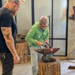 An adult uses a hammer to shape glowing hot metal on an anvil while an ESSA Activity lead volunteer observes in a workshop.