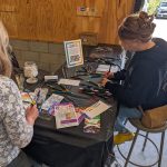 Two participants sit at a table using markers and paint pens to create miniature painting