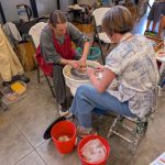 An instructor guides a participant’s hands as they shape a clay piece on a pottery wheel in a studio.