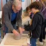 An instructor guides a young participant in shaping a clay coil pot by hand at a workshop table.