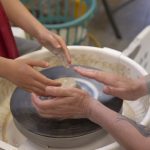 Close-up of an adult guiding a child’s hands as they shape clay on a pottery wheel, with wet clay spinning in a basin.