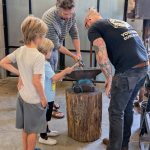 An activity lead assists a child as they hammer heated metal on an anvil during a blacksmithing demonstration.