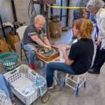 An instructor teaches a participant how to shape clay on a pottery wheel while others observe nearby.