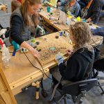 Participants work together at a stained glass station, soldering pieces while wearing protective gloves and eyewear.