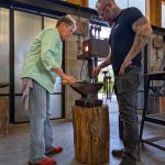 An instructor stands beside a participant as they shape heated metal on an anvil in a blacksmithing studio.