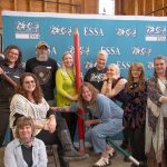 The ESSA Staff pose together in front of an ESSA backdrop, holding oversized art tools including a large red pencil and a hammer.