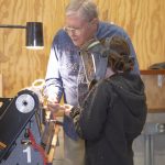 An instructor stands beside a young person wearing a face shield, guiding them as they use a wood lathe covered in wood shavings.