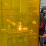 Welding activity viewed through a yellow safety screen as sparks fly inside the workspace.