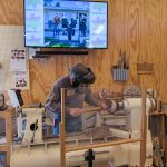 Person wearing a protective face shield uses a wood lathe to shape a spinning piece of wood, with wood shavings scattered across the machine in a workshop. A screen above displays “Hands On ESSA.”