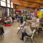 A busy workshop filled with people of all ages participating in hands-on art activities, including pottery and metalwork, inside a large studio with tools and equipment visible throughout.