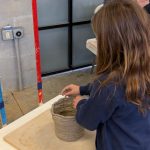 A young person shapes a coil-built clay pot on a worktable, carefully pressing layers of clay together in a studio setting.
