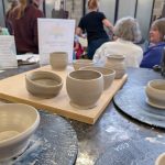 Several freshly thrown clay bowls and vessels sit on pottery wheels and a wooden board, with people working and talking in the background.
