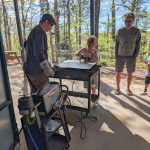 A child wearing gloves works with a tool on a metal surface at an outdoor workstation, while two adults stand nearby watching in a wooded setting.