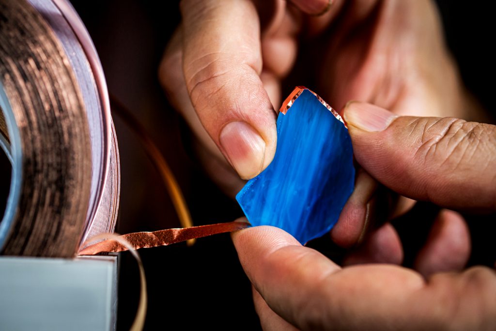 Hands applying copper foil to a piece of blue stained glass