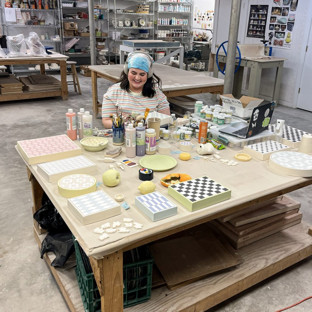 Erin Harris wears headphones at a studio table covered with ceramic food sculptures, patterned display forms, paints, and tools.