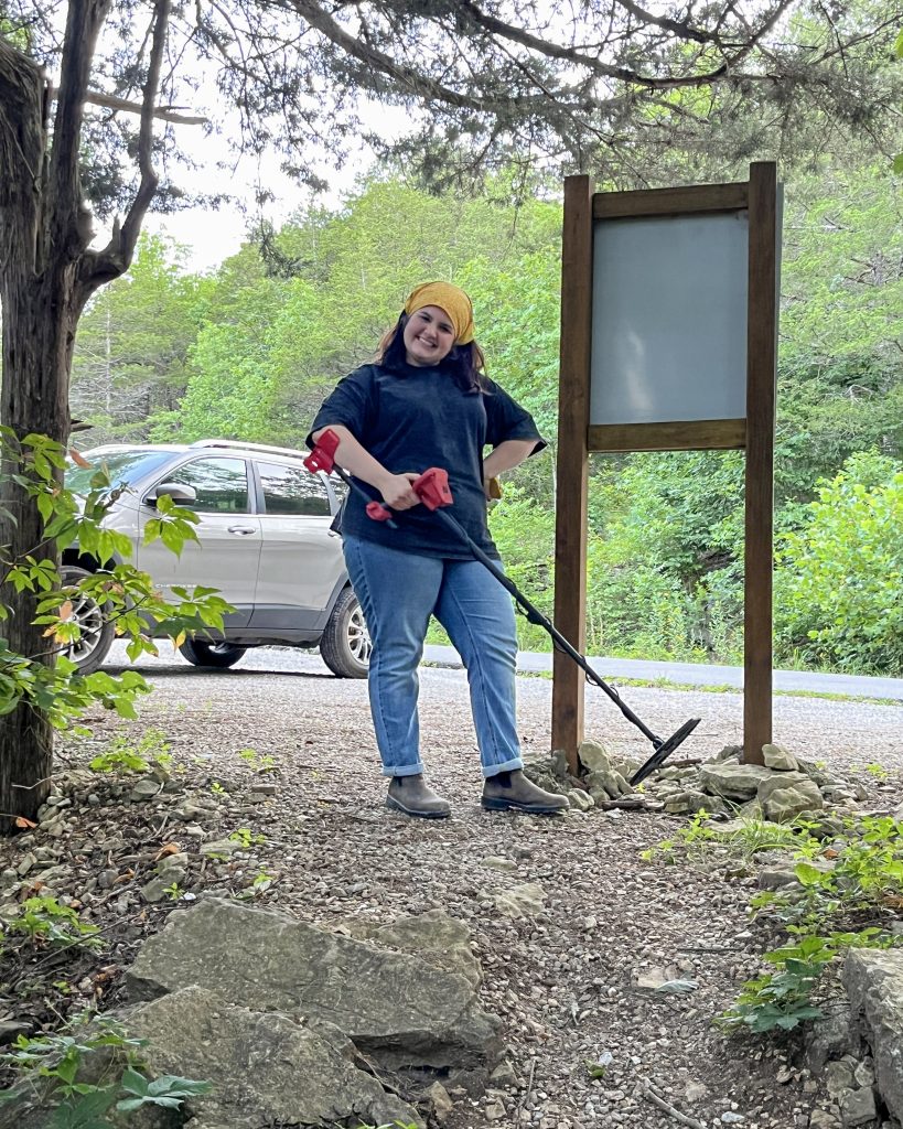 Erin Harris smiles while holding a metal detector in a rocky wooded area in Northwest Arkansas.