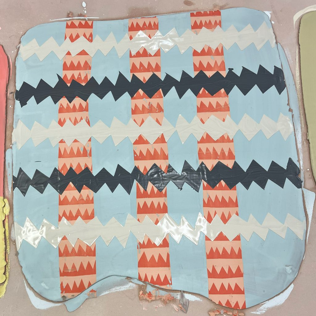 Blue clay slab painted with repeating orange, black, and white patterns before being formed into a ceramic vessel.